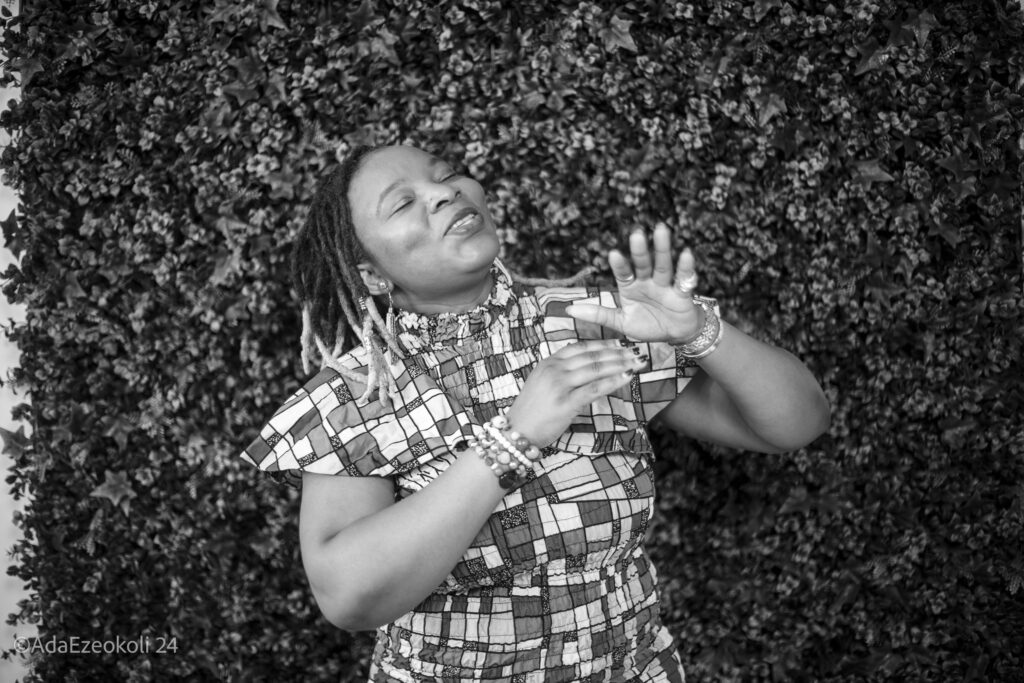 An African woman singing and dancing in front of a wall of green leaves.