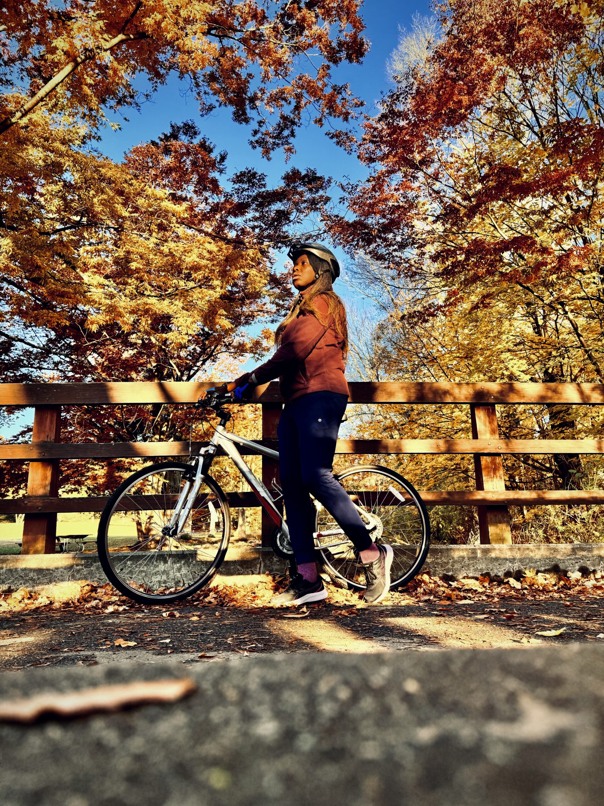A black woman in a helmet walking her bike over a bridge during fall